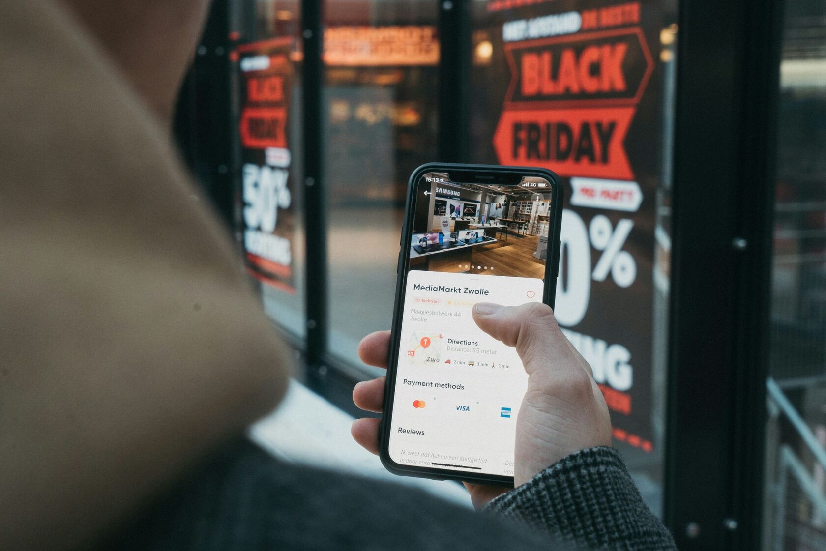 Person browsing store information and payment methods (including Visa and Mastercard) on a smartphone in front of a shop with large &amp;amp;amp;amp;amp;amp;amp;amp;quot;Black Friday 50% Off&amp;amp;amp;amp;amp;amp;amp;amp;quot; sale signs.